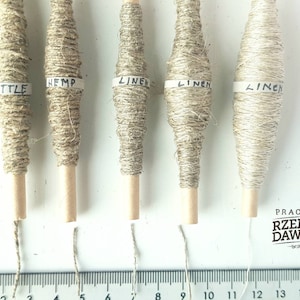 May include: Five spools of natural fiber yarn, labeled "Hemp", "Linen", "Linen", "Linen", and "Nettle". The spools are arranged in a row on a white surface with a ruler underneath.