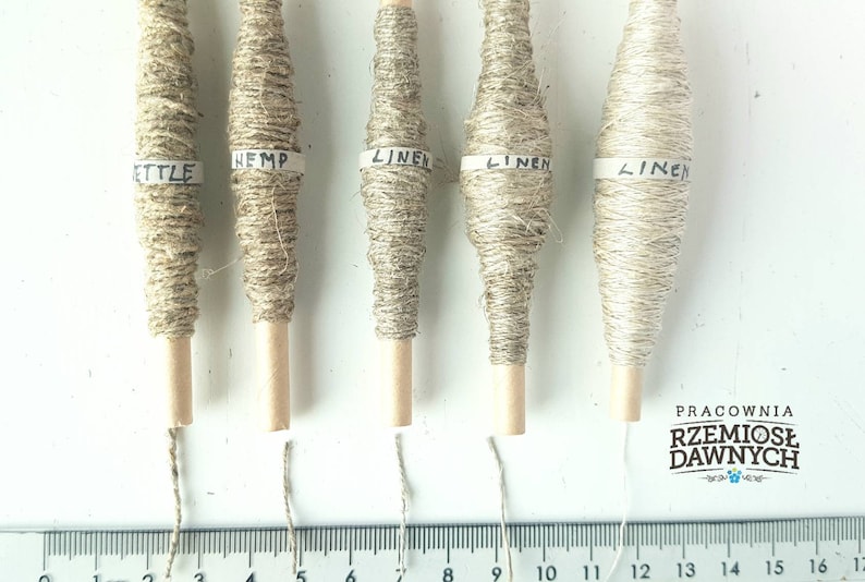 May include: Five spools of natural fiber yarn, labeled "Hemp", "Linen", "Linen", "Linen", and "Nettle". The spools are arranged in a row on a white surface with a ruler underneath.