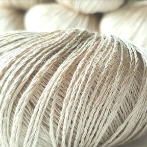 May include: A close-up of a ball of beige yarn. The yarn is made from natural fibres and has a soft, textured appearance.