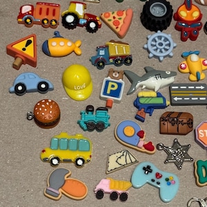 May include: A collection of colorful miniature charms in various shapes, including a train, car, pizza slice, shark, and a yellow hard hat with the word "LOVE". Other charms include a bus, a video game controller, and a treasure chest.