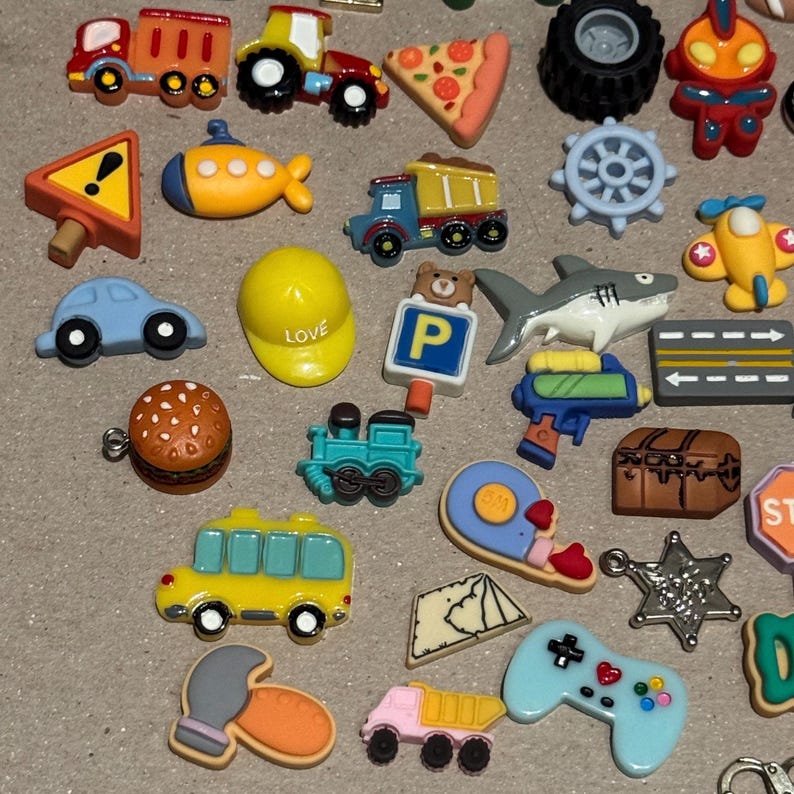 May include: A collection of colorful miniature charms in various shapes, including a train, car, pizza slice, shark, and a yellow hard hat with the word "LOVE". Other charms include a bus, a video game controller, and a treasure chest.
