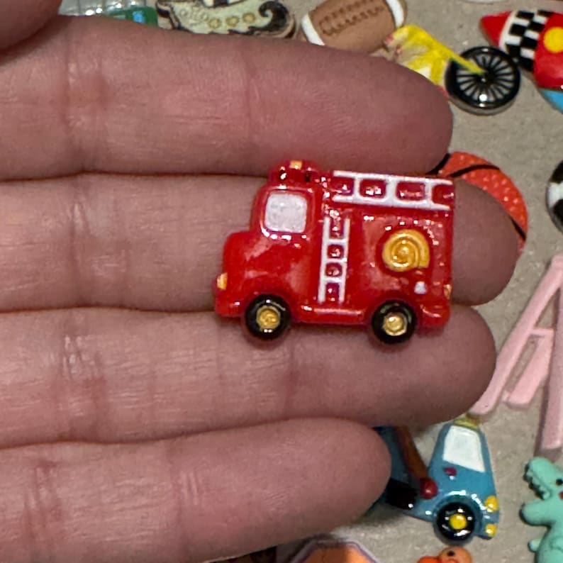 May include: A small, red fire truck charm with black and gold wheels. The miniature vehicle features a white ladder, a gold-colored hose reel, and a white roof. This charm is likely used for crafting or jewelry making.