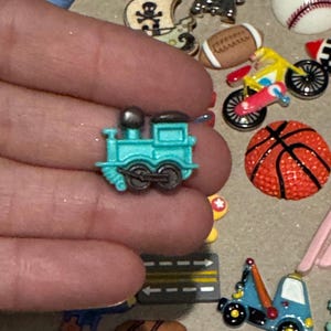 May include: A small, turquoise train charm with black wheels and a silver smokestack. The charm is held in a hand, surrounded by other miniature charms, including a football, a basketball, and a bicycle.