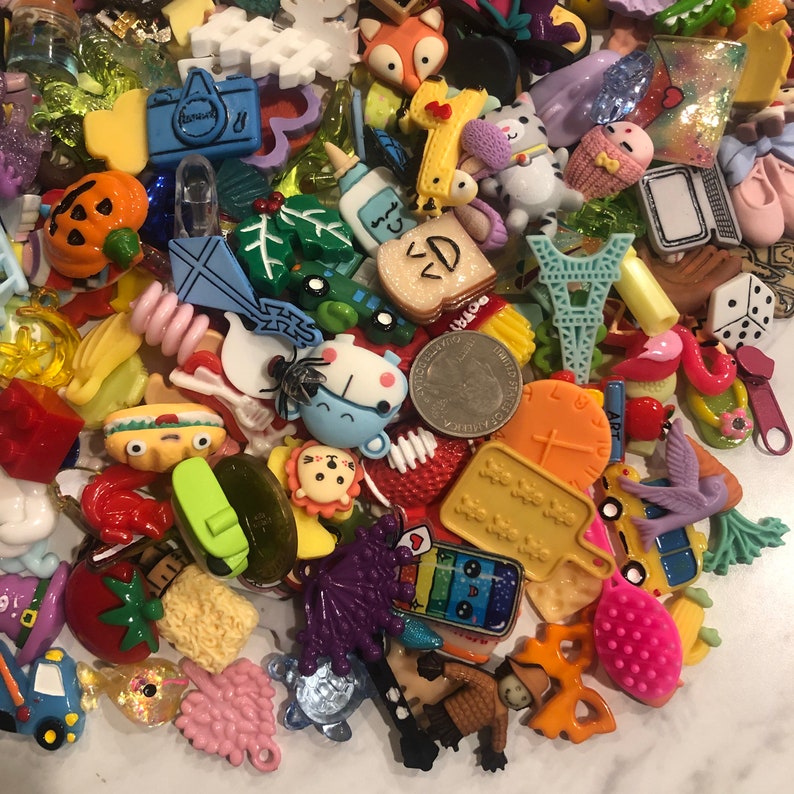May include: A collection of colorful resin charms and embellishments in various shapes, including a camera, a fox, a cat, a pumpkin, a slice of toast, and a miniature Eiffel Tower. The charms are scattered across a white surface, creating a vibrant and playful assortment.