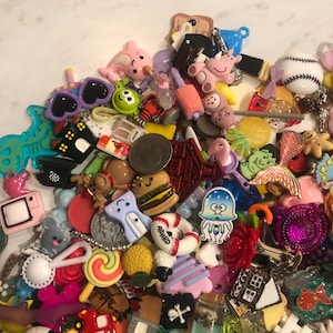 May include: A collection of colorful resin charms and embellishments, including a pink dolphin, sunglasses, a hamburger, a schoolhouse, and a baseball. Other charms include a TV, a lobster, and a mermaid tail. The charms are scattered on a white surface.