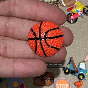 May include: A small, orange basketball charm with black lines. The charm has a textured surface and is held in a hand. Other miniature charms, including a football and a motorcycle, are visible in the background.