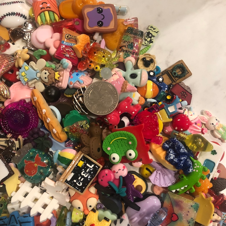 May include: A collection of colorful resin charms in various shapes and sizes, including food items, animals, and other decorative elements. The charms are in shades of pink, green, yellow, and blue, with a quarter coin for size reference.