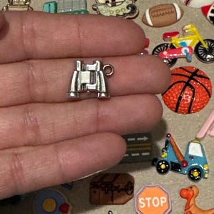May include: A small, silver-colored charm in the shape of binoculars. The charm is held in a hand, with other miniature charms of a football, basketball, and motorcycle visible in the background. The charm is likely for jewelry making or crafting.