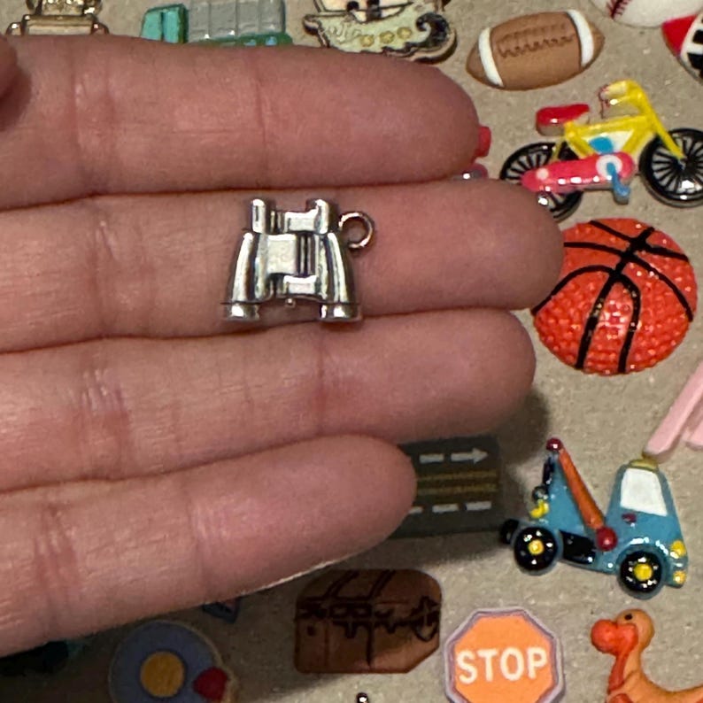May include: A small, silver-colored charm in the shape of binoculars. The charm is held in a hand, with other miniature charms of a football, basketball, and motorcycle visible in the background. The charm is likely for jewelry making or crafting.