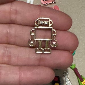 May include: A small, gold-colored robot charm with a rectangular body, rounded arms and legs, and a detailed head. The charm is held in a hand, with other small charms visible in the background.
