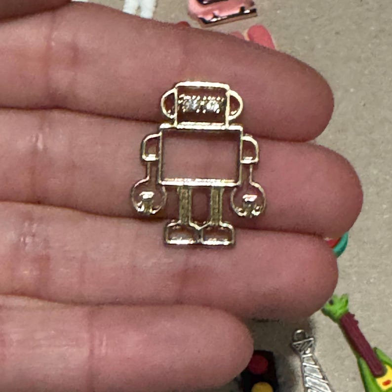 May include: A small, gold-colored robot charm with a rectangular body, rounded arms and legs, and a detailed head. The charm is held in a hand, with other small charms visible in the background.