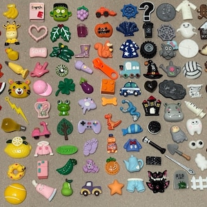 May include: A collection of colorful resin embellishments in various shapes, including food, animals, and objects. The assortment features a variety of colors and designs, such as a red apple, a yellow banana, and a green frog. The word "English" is visible on one of the embellishments.