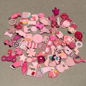 May include: A collection of pink charms and embellishments, including a flamingo, a shoe, a book with the word "English", a crown, and a tennis racket. Other shapes include stars, hearts, and a clock. The charms are scattered on a gray surface.