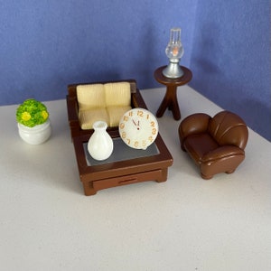 May include: A miniature living room set with a brown couch, a brown armchair, a coffee table with a clock and a white vase, a small table with a lamp, and a potted plant with green leaves.