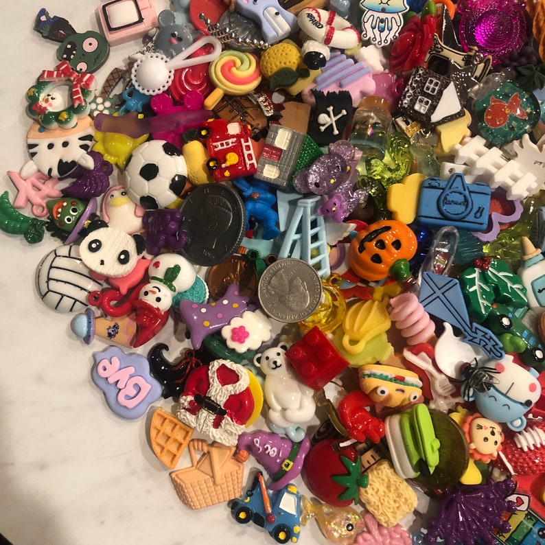 May include: A collection of colorful resin charms in various shapes, including a soccer ball, panda, snowman, and a pumpkin. Other charms include a camera, a ladder, and a waffle. The charms are in a variety of colors, including red, blue, green, and yellow.