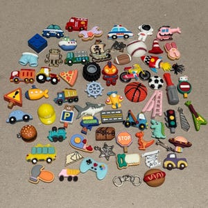 May include: A collection of colorful, miniature decorative charms in various shapes. The charms include a fire truck, a baseball, a dinosaur, a stop sign, a hamburger, a video game controller, and a basketball. The charms are made of plastic.