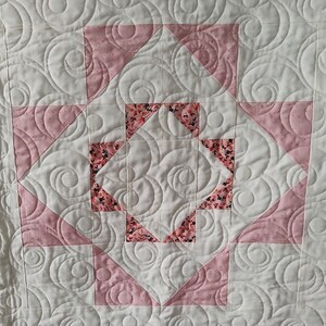 May include: A white quilt with a pink and white geometric pattern. The quilt is quilted with a swirling pattern.