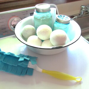 May include: A white bowl filled with white dryer balls, two blue glass jars with lids, and a blue and yellow cleaning brush. The bowl is sitting on a white worktop next to a stainless steel sink.