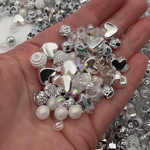 May include: A collection of silver and white beads, including hearts, stars, and round beads. The beads are in a variety of sizes and shapes.