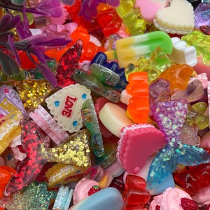 May include: A colourful collection of resin charms in diverse shapes, such as hearts, bows, mermaid tails, and food items like ice cream and cakes. The charms are in shades of pink, red, orange, yellow, green, and blue, with glitter accents and the word "Love" on one charm.