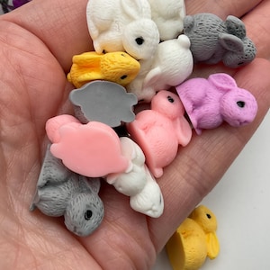 May include: A collection of small, colourful bunny figurines made of resin. The bunnies are in various poses and colours, including pink, purple, yellow, and grey.