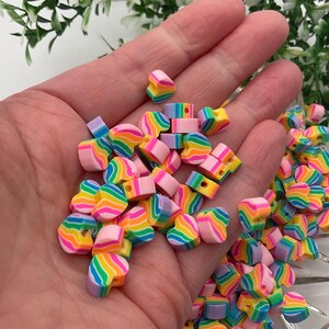 May include: A collection of colourful rainbow heart-shaped beads with a hole in the centre. The beads are made of polymer clay and are perfect for jewellery making or crafting.