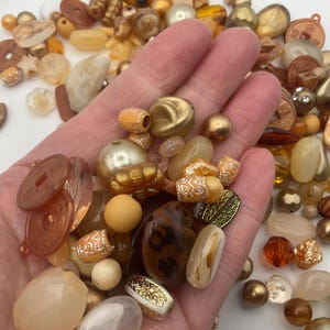 May include: A collection of gold, brown, and clear beads in various shapes and sizes. The beads are made of glass, plastic, and metal. Some of the beads have intricate designs.