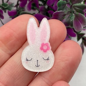 May include: White glitter bunny with pink glitter ears and a pink flower on its head. The bunny has closed eyes and a small black nose.
