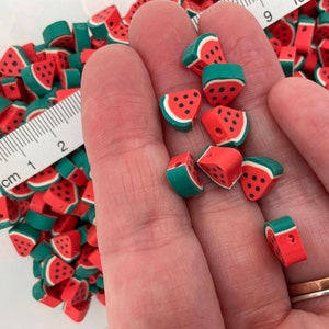 May include: A close-up of a hand holding a pile of red and green watermelon-shaped beads. The beads are made of polymer clay and have a realistic watermelon design.