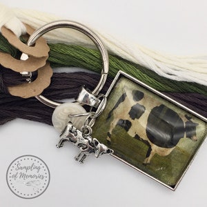 Könnte beinhalten: Ein silberner Schlüsselanhänger mit einem Anhänger einer Kuh auf einer grünen Weide. Der Anhänger ist in einem silbernen Rahmen. Der Schlüsselanhänger hat auch einen kleinen silbernen Kuh-Anhänger und einen weißen Knopf.