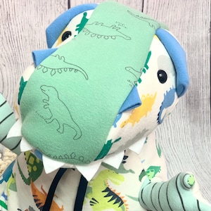 May include: A stuffed dinosaur toy with a green and blue color scheme. The toy features a green head covering with dinosaur outlines, blue accents, and a white body with various dinosaur patterns. It has a white collar and a striped arm.