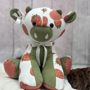 May include: A plush toy cow with a white, green, and pink fabric. The cow has a strawberry pattern and large black eyes. The cow is sitting on a white blanket.