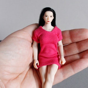 K&ouml;nnte beinhalten: Eine kleine Puppe, die ein rosa Kleid tr&auml;gt, schwarze Haare hat und einen neutralen Gesichtsausdruck zeigt. Die Puppe sitzt in der Handfl&auml;che einer Hand.