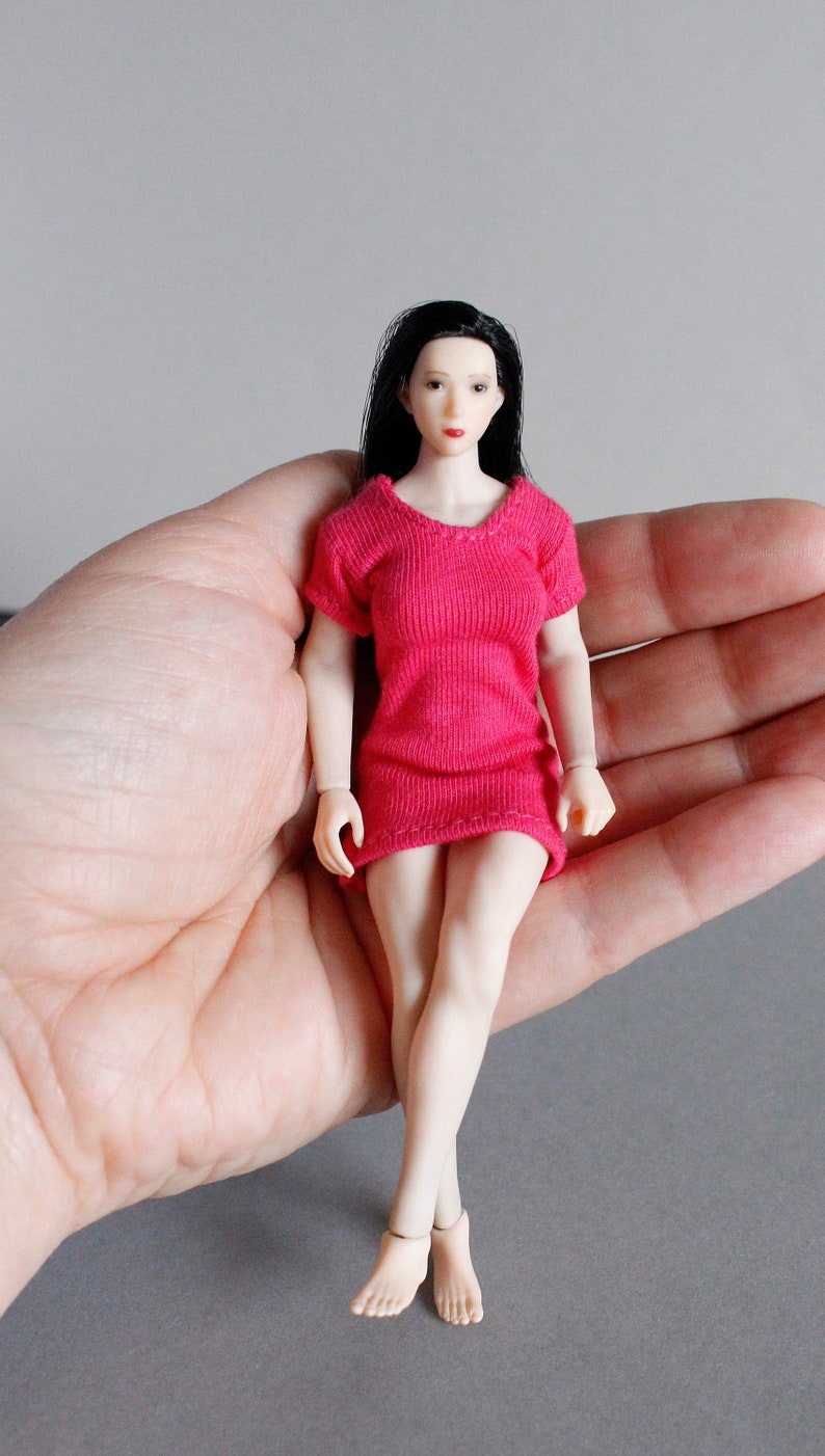 K&ouml;nnte beinhalten: Eine kleine Puppe, die ein rosa Kleid tr&auml;gt, schwarze Haare hat und einen neutralen Gesichtsausdruck zeigt. Die Puppe sitzt in der Handfl&auml;che einer Hand.