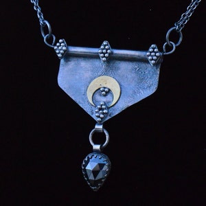 Könnte beinhalten: Silberfarbene Halskette mit Anhänger. Der Anhänger hat ein strukturiertes, schildförmiges Element, einen goldenen Halbmond und einen tropfenförmigen schwarzen Edelstein. Die Kette hat eine feine Kette und Perlenakzente. Der Anhänger ist ca. 5 cm lang.