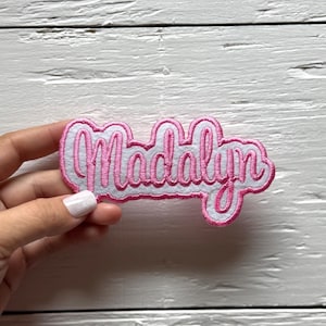 May include: A pink and white embroidered patch with the name "Madalyn" in a cursive font. The patch has a white border and is likely designed to be sewn or ironed onto fabric. The background is a light wood surface.