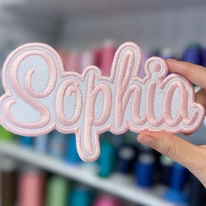 May include: A custom embroidered patch in a cursive font spelling "Sophia". The patch features a light pink embroidered name with a white border. The patch is held in a hand, with a blurred background of colorful spools.