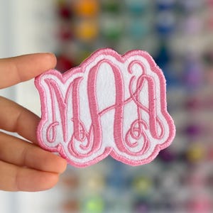 May include: A pink and white embroidered monogram patch featuring the letters "MAA" in a decorative script font. The patch has a scalloped edge and is held up by a hand.