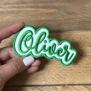 May include: Embroidered patch in the shape of the name "Oliver" in a cursive font. The name is in white with a green outline and border. The patch is held by a hand against a wooden background.
