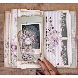 May include: An open scrapbook with a vintage aesthetic. The pages feature floral and clock designs in shades of pink, purple, and cream. A decorative tag with a clock and roses hangs from the frame. The scrapbook is on a wooden surface.