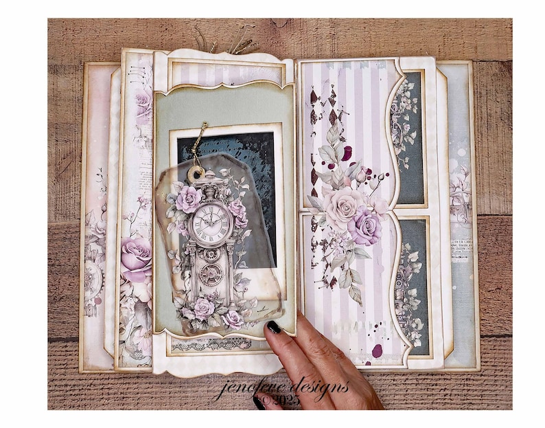 May include: An open scrapbook with a vintage aesthetic. The pages feature floral and clock designs in shades of pink, purple, and cream. A decorative tag with a clock and roses hangs from the frame. The scrapbook is on a wooden surface.