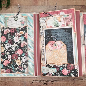 May include: Open scrapbook with floral and striped patterned pages. The left side features a floral print pocket and a tag with a floral design. The right side has a tag that says "adore you". The scrapbook is open on a wooden surface.