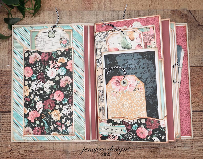 May include: Open scrapbook with floral and striped patterned pages. The left side features a floral print pocket and a tag with a floral design. The right side has a tag that says "adore you". The scrapbook is open on a wooden surface.