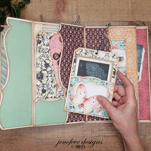 May include: A handmade scrapbook album with a butterfly design. The album features a variety of patterned papers in shades of pink, green, and cream. The album is open, revealing several pages and pockets, with a hand holding a tag with a butterfly illustration. The text "jenofeve designs &copy;2025" is visible.