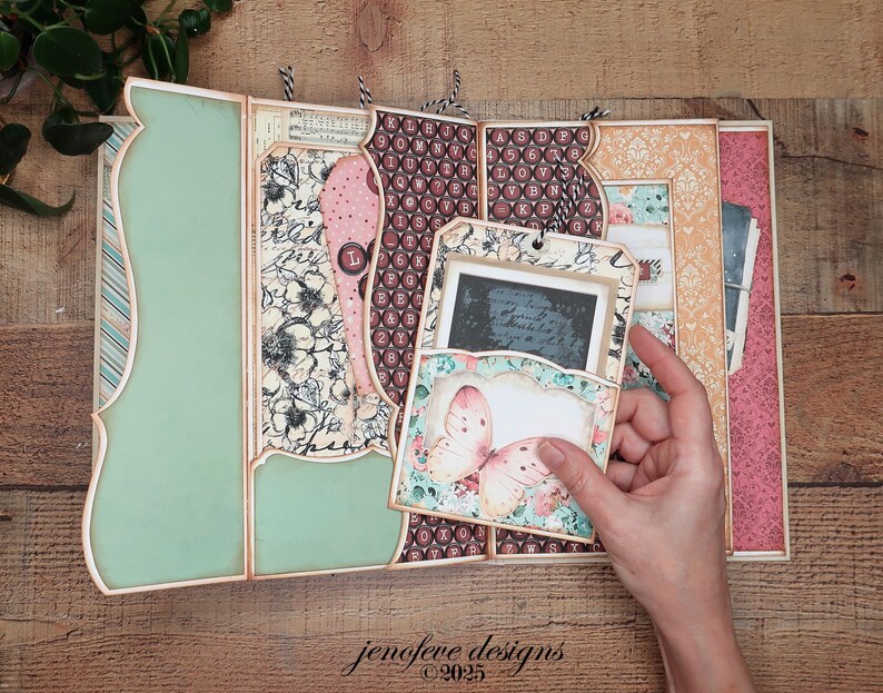 May include: A handmade scrapbook album with a butterfly design. The album features a variety of patterned papers in shades of pink, green, and cream. The album is open, revealing several pages and pockets, with a hand holding a tag with a butterfly illustration. The text "jenofeve designs &copy;2025" is visible.