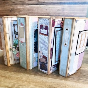 May include: Six decorative scrapbook albums with a vintage theme. The albums are made of cardboard and have a variety of embellishments, including paper flowers, lace, and ribbons. The albums are closed and are arranged in a row on a wooden surface. The albums have a variety of colors, including brown, blue, green, and pink. The albums have text on the covers, including 'love' and '2019'. 