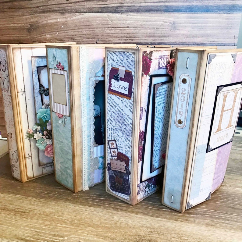 May include: Six decorative scrapbook albums with a vintage theme. The albums are made of cardboard and have a variety of embellishments, including paper flowers, lace, and ribbons. The albums are closed and are arranged in a row on a wooden surface. The albums have a variety of colors, including brown, blue, green, and pink. The albums have text on the covers, including 'love' and '2019'. 