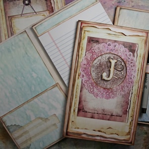 May include: A vintage-style journal with a distressed brown cover and a decorative pink and white lace design. The journal has a large letter "J" in the center of the lace design. The journal is open to a blank page with lined paper.