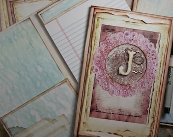 May include: A vintage-style journal with a distressed brown cover and a decorative pink and white lace design. The journal has a large letter "J" in the center of the lace design. The journal is open to a blank page with lined paper.
