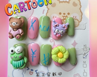 Può includere: Un set di dieci unghie finte verdi e rosa con disegni di animali e fiori dei cartoni animati. Le unghie hanno disegni dipinti a mano e includono un orso, una rana, un fiore e un coniglio. Le unghie sono esposte su una carta bianca con la parola "Cartoon" in lettere colorate.